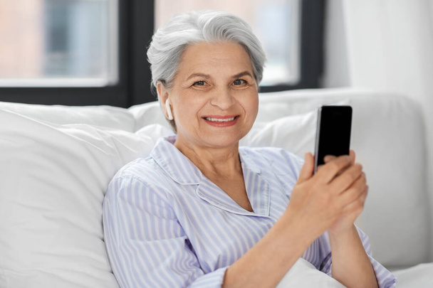 senior woman with smartphone and earphones in bed - Foto, Imagen
