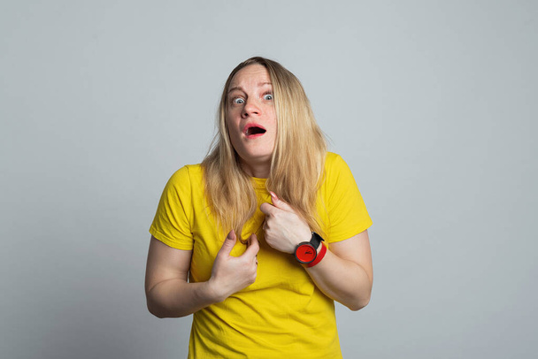 Unzufriedene Frau zeigt empört auf sich selbst, Unzufriedenheit über ungerechtfertigte Behauptung, fragt, warum ich, gelbes T-Shirt tragend, über grauem Hintergrund stehe - Foto, Bild