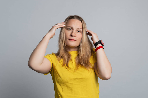 Oh, no. Photo of serious woman looking at camera, thinking and touching her temples, wearing casual yellow t shirt. Studio shot, gray background - Photo, Image