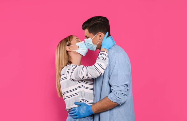 Paar in medische maskers en handschoenen proberen te kussen op roze achtergrond - Foto, afbeelding