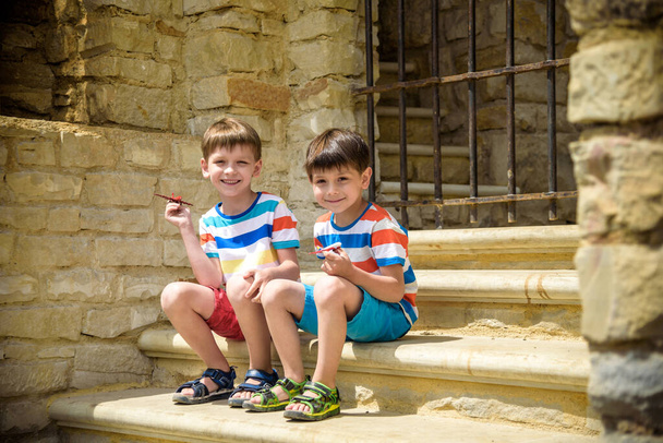 The children playing on the ruins of ancient building with metal gate an archaeological site of an ancient city. Two boys sitting and play with toy aircraft plane. Travel concept. - Photo, Image