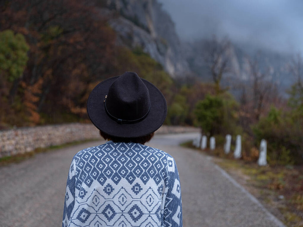Vue arrière de la femme hipster élégant chapeau et poncho marchant sur une route de montagne. Le concept de voyage et d'errance. Un moment atmosphérique incroyable - Photo, image