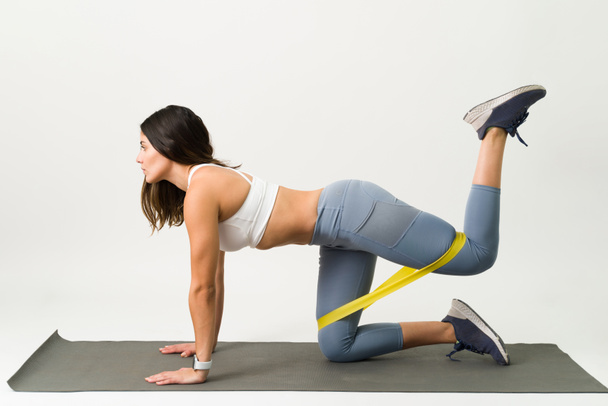 Attractive young woman in activewear working out on an exercise mat and doing kickback exercises with a resistance band - Photo, Image