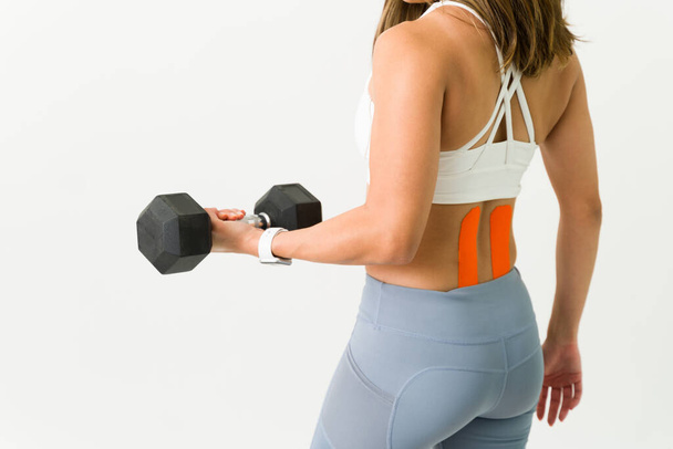 Fit female athlete seen from behind lifting a dumbbell weight and working out her strong arms. Young sporty woman using kinesiology tape on her lower back - Photo, Image