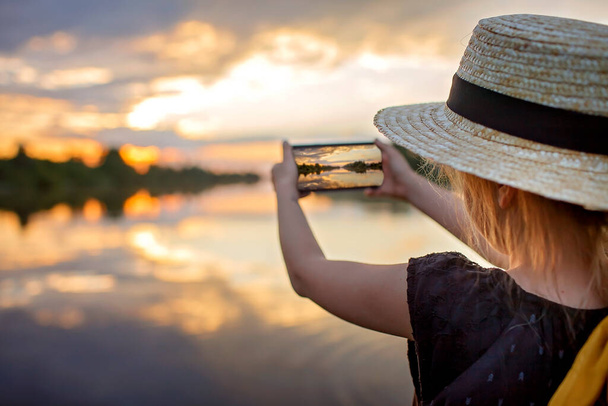 Yazın hasır şapkalı kız gölde gün batımında cep telefonu için selfie çekiyor, yaşam tarzı, yerel seyahat. - Fotoğraf, Görsel
