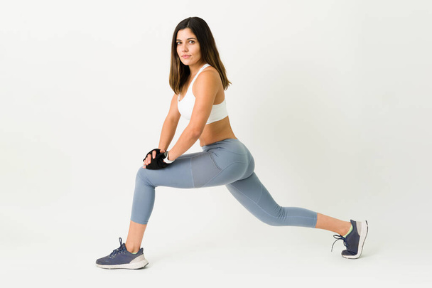Retrato de uma jovem bonita em roupas esportivas fazendo contato visual enquanto faz exercícios lunges contra um fundo branco - Foto, Imagem