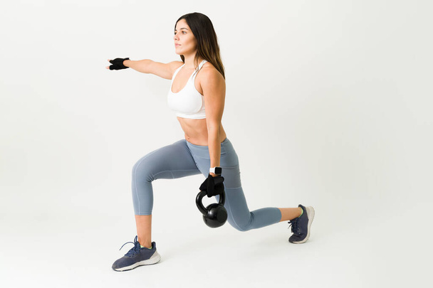 Beautiful sporty woman doing lunges while holding a kettlebell. Fit strong athlete exercising with weights for her daily training program - Photo, Image