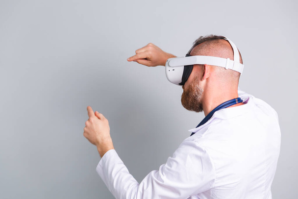 Handsome male doctor on gray background in virtual reality glasses back view touching air - Foto, Imagem