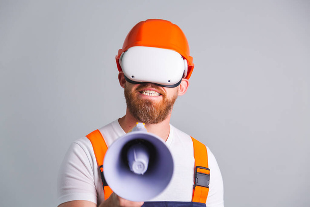 Pohledný muž inženýr v budování ochranné přilby na šedém pozadí křičí v megafonu ve vr brýle - Fotografie, Obrázek