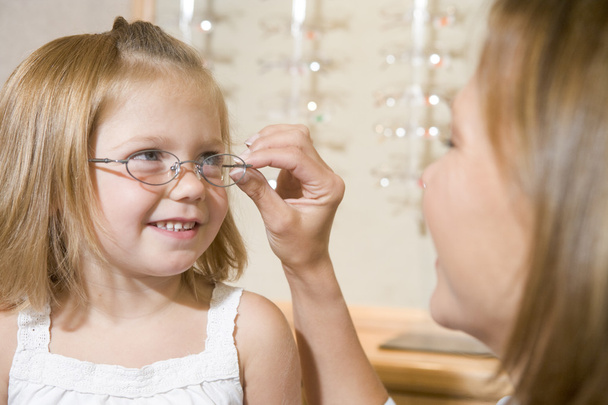 Woman trying eyeglasses on young girl at optometrists smiling - Φωτογραφία, εικόνα