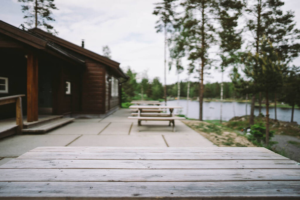Outdoor wooden table in front if a cottage with a lake in the background and high trees - Фото, зображення