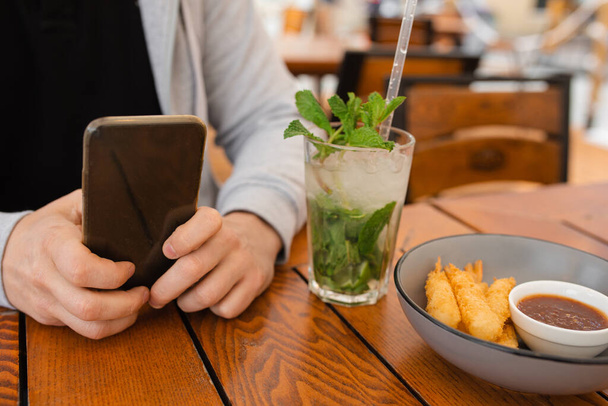 Κοντινό πλάνο Φωτογραφία των χεριών του ανθρώπου στο τραπεζάκι του καφέ χρησιμοποιώντας smartphone δίπλα στο cooling mojito cocktail, mobile device concept - Φωτογραφία, εικόνα