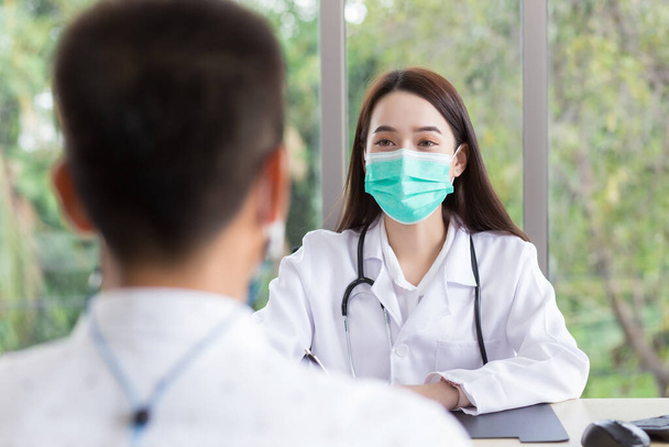 Asiática profesional médico femenino lleva abrigo médico y mascarilla mientras examina y habla con un hombre paciente en la sala de examen en el hospital. - Foto, Imagen