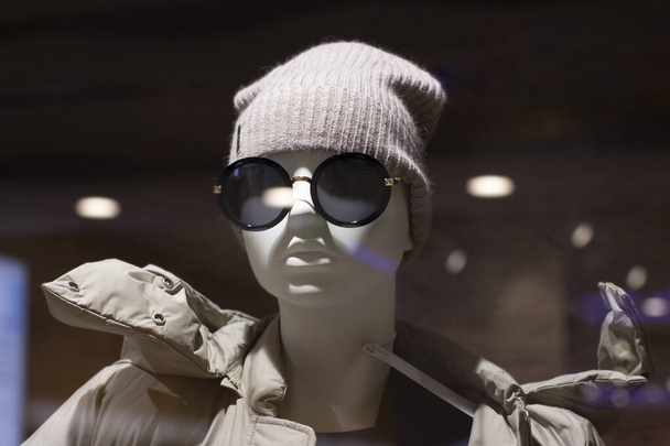 La tête d'un mannequin dans un chapeau et des lunettes de soleil, les reflets du verre et des lampes. Mannequin en bonnet tricoté et manteau close-up - Photo, image