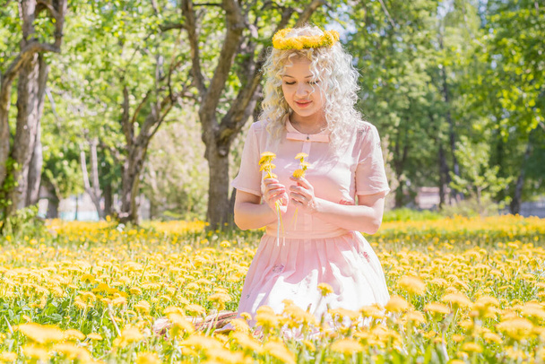 A happy young girl in a pink dress and with a wreath of yellow dandelions on her head sits on the lawn and looks at the flowers in her hands. A bright sunny day. - Valokuva, kuva