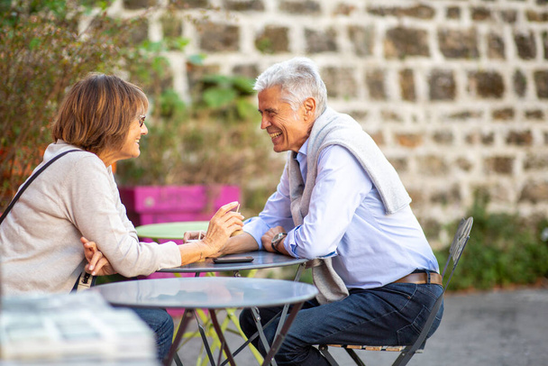 Zijportret ouder paar zitten aan tafel buiten glimlachend naar elkaar - Foto, afbeelding
