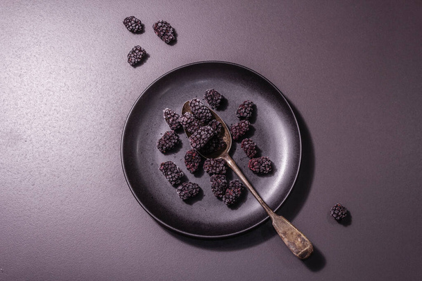 Βατόμουρα σε κεραμικό πιάτο. Κατεψυγμένα φρούτα για υγιεινή διατροφή. Minimalistic concept, μαύρη πέτρα, σκληρό φως, σκούρα σκιά, top view - Φωτογραφία, εικόνα