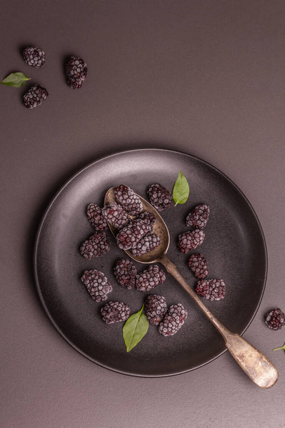 Des mûres sur une assiette en céramique. Fruits surgelés pour une alimentation saine. Concept minimaliste, fond en béton de pierre noire, vue de dessus - Photo, image