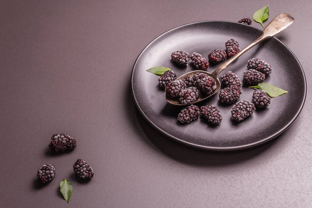 Blackberries on a ceramic plate. Frozen fruits for a healthy food diet. Minimalistic concept, black stone concrete background, hard light, dark shadow, flat lay - 写真・画像