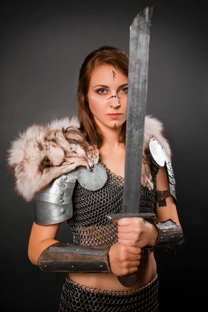 Medieval woman warrior in chain mail armor and polar fox fur on her shoulders standing with a sword in hand on a dark background. Fantasy barbarian, viking. - 写真・画像