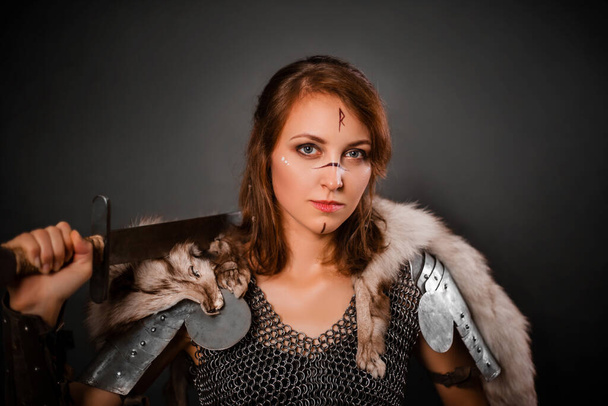 Medieval woman warrior in chain mail armor and polar fox fur on her shoulders standing with a sword in hand on a dark background. Fantasy barbarian, viking. - Foto, imagen