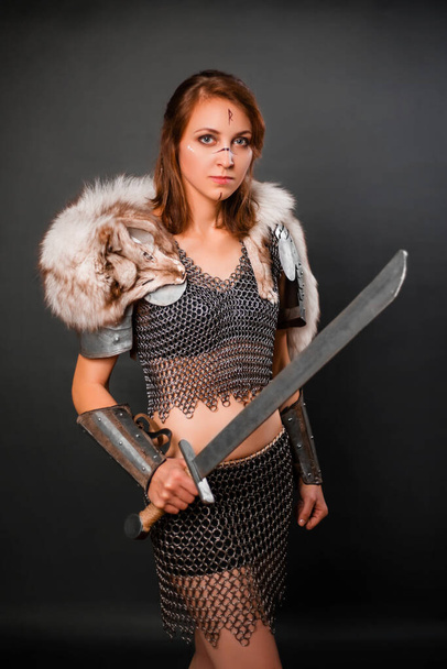 Medieval woman warrior in chain mail armor and polar fox fur on her shoulders standing with a sword in hand on a dark background. Fantasy barbarian, viking. - Foto, Bild