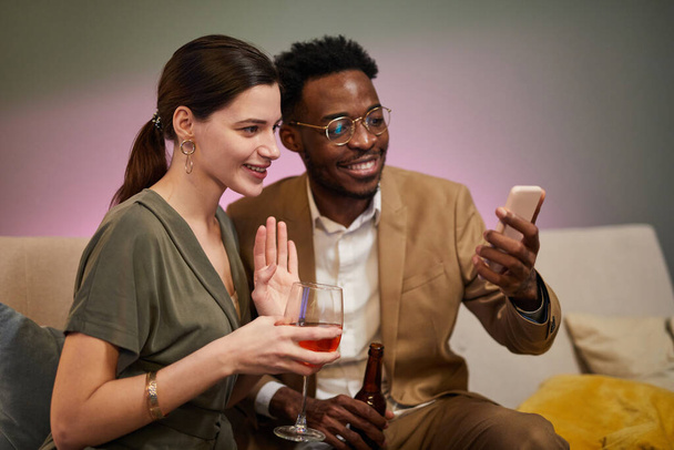 家の設定でビデオチャットを介してオンラインでお祝いしながら、笑顔のカップルの肖像画は、スマートフォンの画面に手を振る - 写真・画像