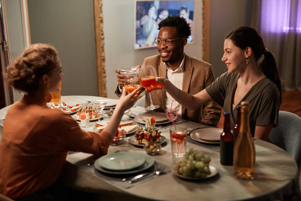 Multietnická skupina přátel si užívá večerní večeři v interiéru a cinkání sklenic nad slavnostním stolem - Fotografie, Obrázek