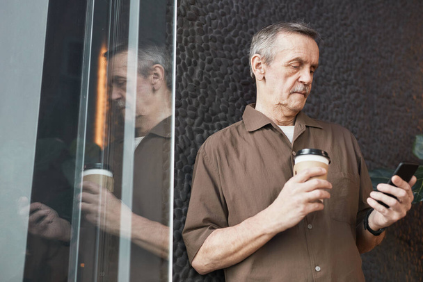 Abaixo vista do gerente caucasiano sênior ocupado sério com café takeout em pé na parede preta e mensagem de leitura no telefone - Foto, Imagem
