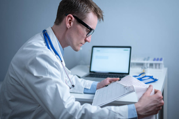 Jeune beau médecin homme en lunettes et blouse blanche examine les résultats de l'examen électrocardiographique du cœur du patient dans le bureau de l'hôpital. Cardiologue regardant cardiogramme au cabinet médical. - Photo, image