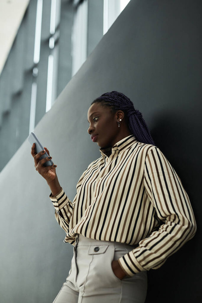 Grafisch zijaanzicht portret van een zelfverzekerde Afrikaans-Amerikaanse zakenvrouw met smartphone terwijl ze tegen een zwarte achtergrond staat - Foto, afbeelding