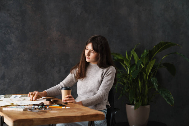 Focused beautiful calligrapher girl drinking coffee while working at table indoors - Foto, imagen