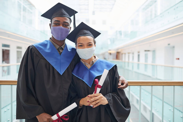Retrato de cintura hacia arriba de dos jóvenes afroamericanos que llevan vestidos de graduación y máscaras mientras posan en interiores - Foto, Imagen