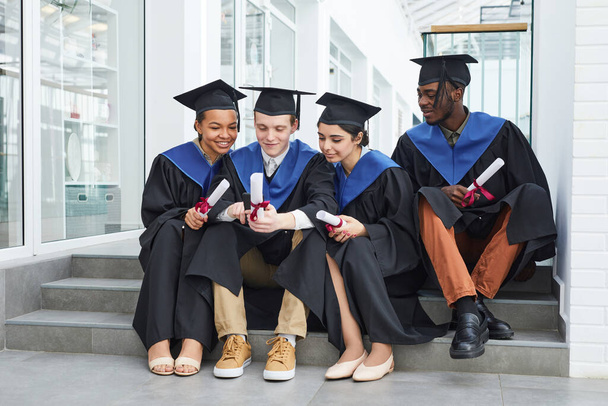 Diverse group of happy young people wearing graduation gowns and using smartphone while sitting on steps outdoors - Foto, afbeelding