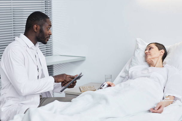 Médico africano recetando un medicamento a una mujer madura mientras está acostada en la cama en el hospital - Foto, Imagen
