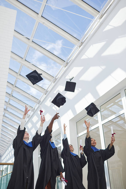 Vertikaler Blick auf eine vielfältige Gruppe von Hochschulabsolventen, die mit Sonnenlicht Hüte in die Luft werfen, Kopierraum - Foto, Bild