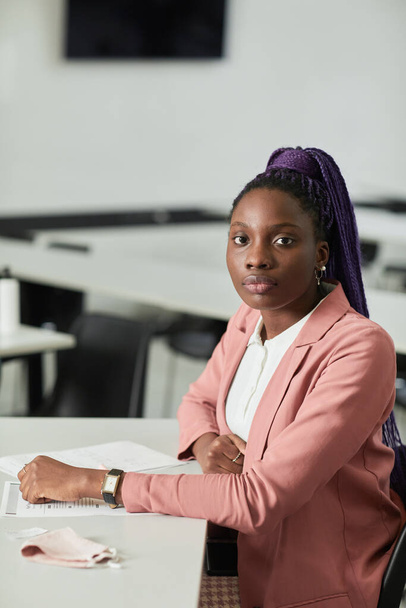 Retrato vertical mínimo de una joven afroamericana mirando a la cámara en la escuela mientras está sentada en el escritorio en el aula - Foto, Imagen