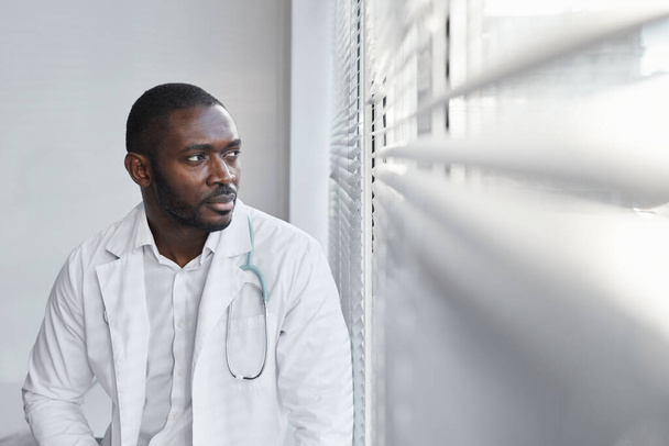 Αφρικανός νεαρός γιατρός κάθεται κοντά στο παράθυρο του νοσοκομείου και βλέπει - Φωτογραφία, εικόνα