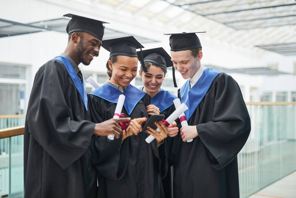 Разнообразная группа счастливых молодых людей, одетых в выпускные платья и использующих смартфон, стоя в помещении в интерьере колледжа - Фото, изображение