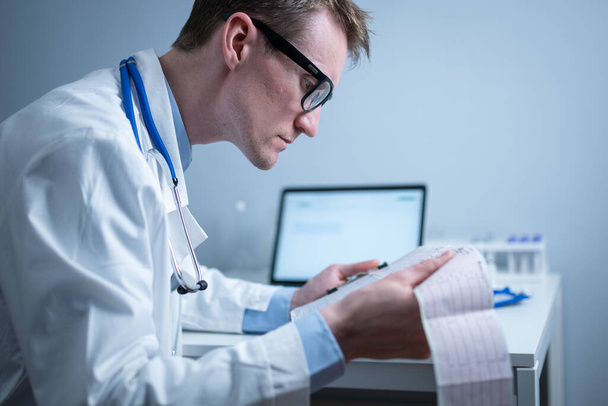 Jeune beau médecin homme en lunettes et blouse blanche examine les résultats de l'examen électrocardiographique du cœur du patient dans le bureau de l'hôpital. Cardiologue regardant cardiogramme au cabinet médical. - Photo, image