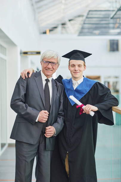 Portrait vertical d'un jeune homme souriant posant avec son père pendant la cérémonie de remise des diplômes à l'intérieur - Photo, image