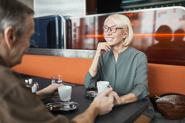 Smiling attractive senior Caucasian woman in eyeglasses sitting at table in bistro cafe and holding hand of boyfriend while listening to him - Photo, Image