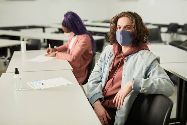 Portret van een langharige jongeman met een masker op op school en kijkend naar de camera terwijl hij achter het bureau in de klas zat, kopieer ruimte - Foto, afbeelding