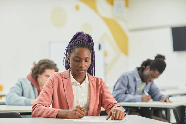 Monipuolinen ryhmä oppilaita, jotka opiskelevat koulussa keskittyen nuoreen afroamerikkalaiseen naiseen, joka istuu pöydän ääressä, kopioi tilaa - Valokuva, kuva