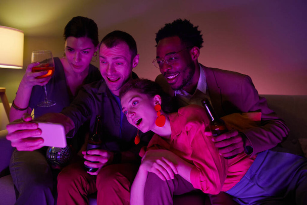 Diversi gruppi di amici che scattano selfie o video chattano insieme durante una festa al coperto illuminata da luci viola - Foto, immagini
