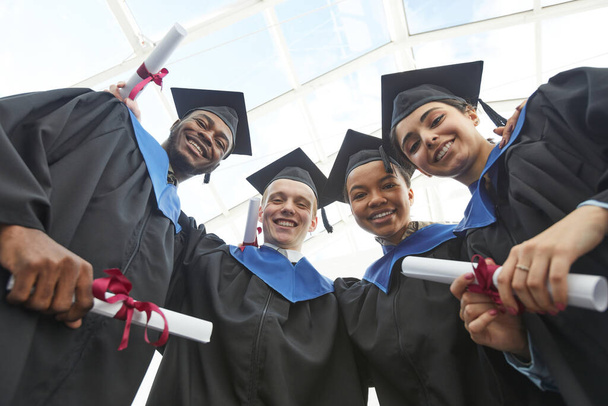 Low angle view at diverse group of happy young people wearing graduation gowns and holding diplomas while looking down at camera - Photo, Image