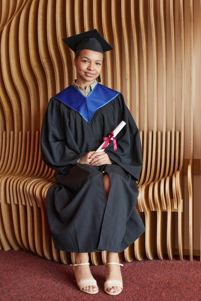 Vertical full length portrait of smiling young woman wearing graduation gown and smiling at camera while posing indoors - Photo, Image