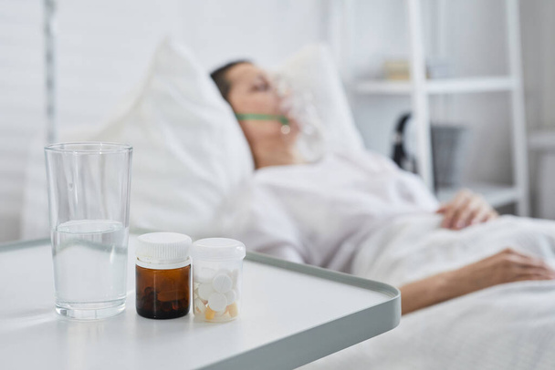 Detailní záběr léků a sklenice vody na stole s nemocným pacientem v pozadí - Fotografie, Obrázek