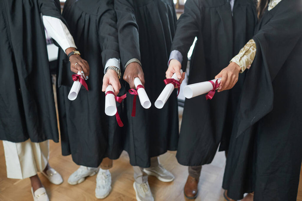 Wysoki kąt widzenia na wieloetniczną grupę młodych ludzi noszących szaty dyplomowe i pokazujących dyplomy przed kamerą - Zdjęcie, obraz