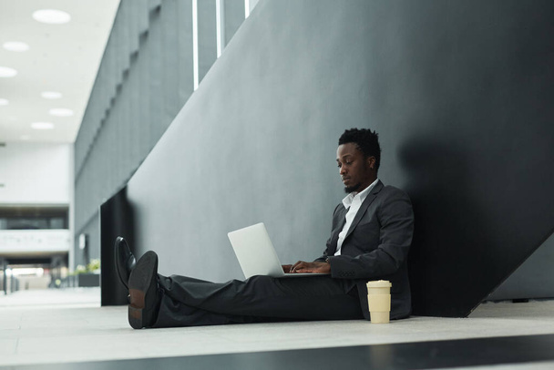 Retrato gráfico de larga duración de un hombre de negocios afroamericano que usa una computadora portátil mientras está sentado en el piso del salón de oficinas, espacio para copiar - Foto, imagen